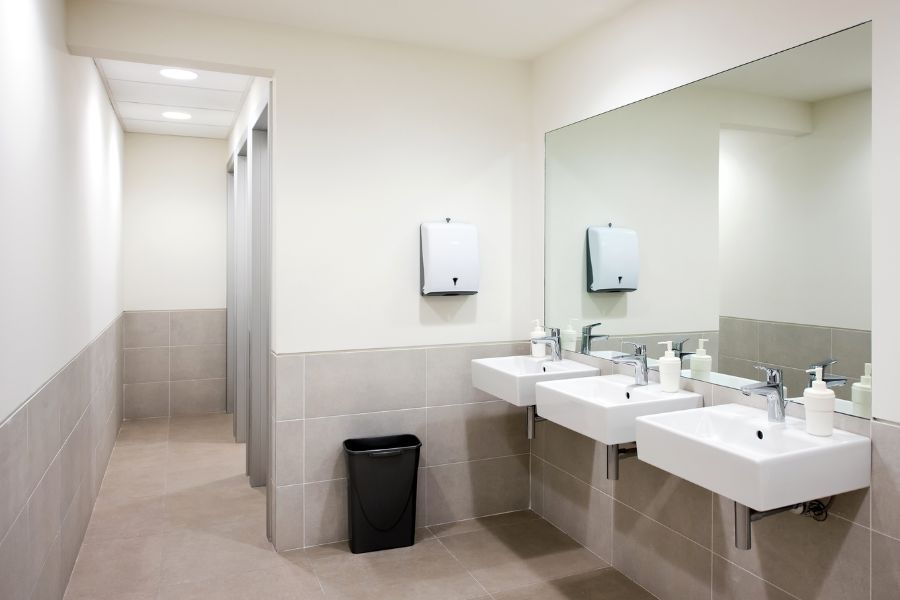A photo of a light coloured commercial washroom. with 2 sinks and mirrors with a. tiled floor.