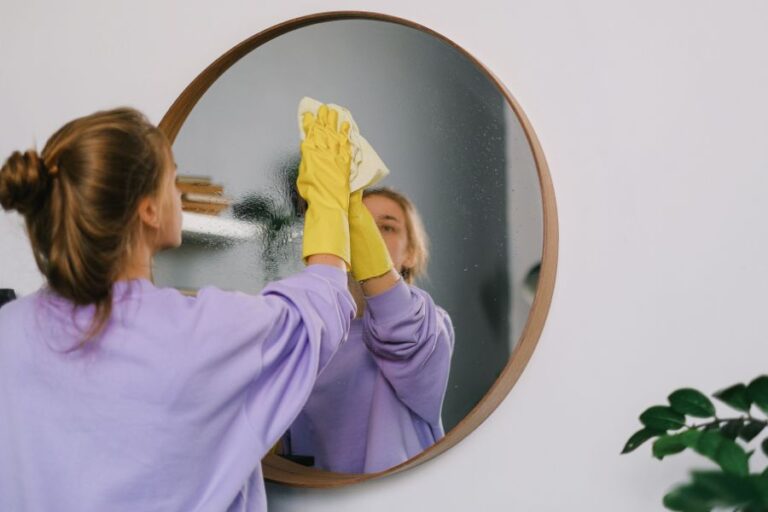 a woman in purple and wearing yellow rubber gloves, cleaning a dirty bathroom mirror
